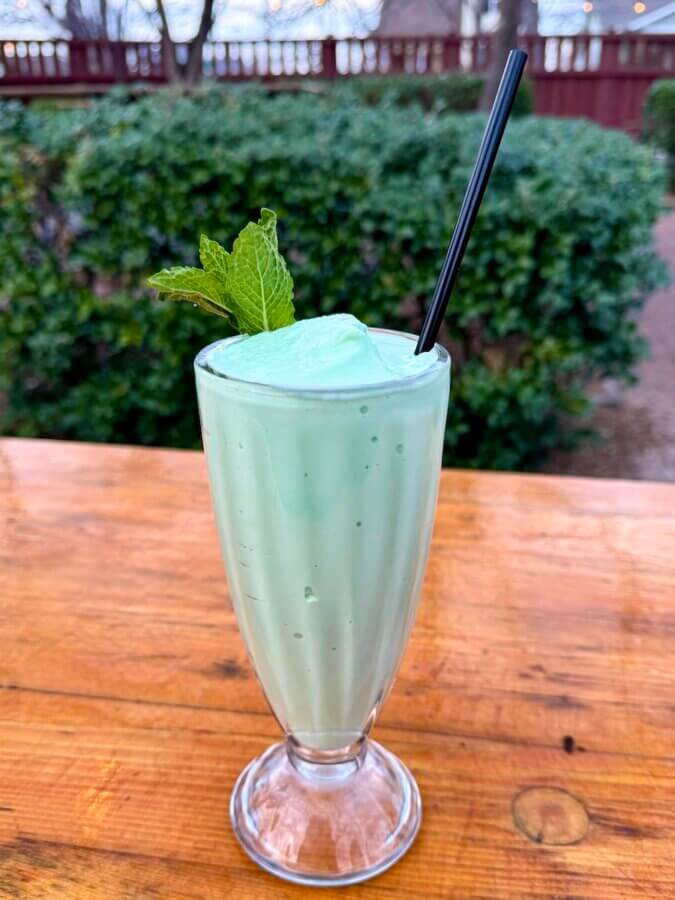 Nashville Steals, Deals & Announcements: March 2026 - 7 A tall glass of light green mint milkshake with a black straw and mint garnish sits on a wooden table outdoors, surrounded by greenery—a refreshing treat perfect for celebrating Nashville Deals.