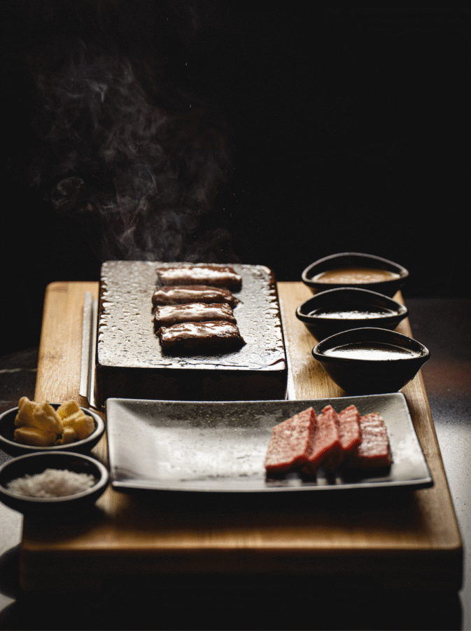 Nashville Steals, Deals & Announcements: March 2026 - 9 Sliced meat grilling on a hot stone with three dipping sauces, lemon wedges, and salt on a wooden board beside raw meat slices on a plate—don’t miss these delicious Nashville deals for your next meal!.