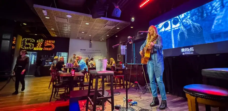 A musician plays guitar and sings on stage in a bar, with people seated at tables and a large screen displaying her image in the background—a lively spot featured in the BNA Dining Guide.
