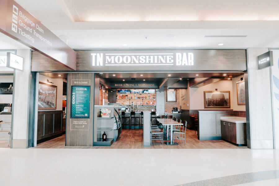Front view of TN Moonshine Bar in an airport, featured in the BNA Dining Guide, with tables, chairs, a menu board, and a bar counter displaying drinks.