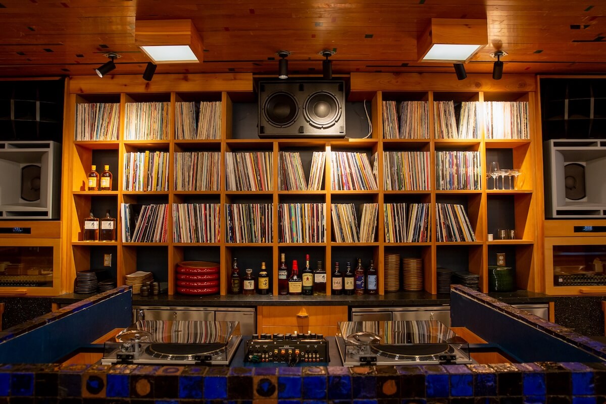 A wooden shelving unit houses vinyl records, speakers, liquor bottles, glasses, and plates—creating the perfect vibe for vinyl lounge spots—with DJ equipment and turntables set up on a counter in the foreground.