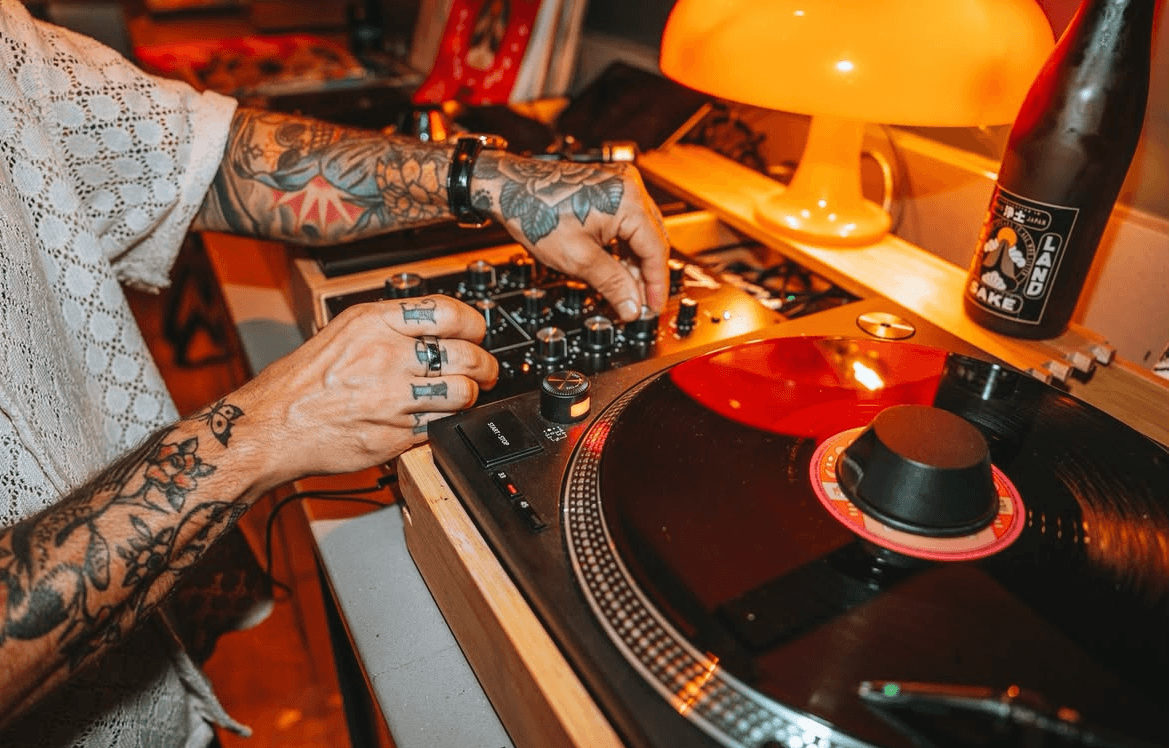 Tattooed person adjusts controls on a DJ mixer next to a turntable, creating vibrant energy in a vinyl lounge, with an orange lamp and a bottle in the background.