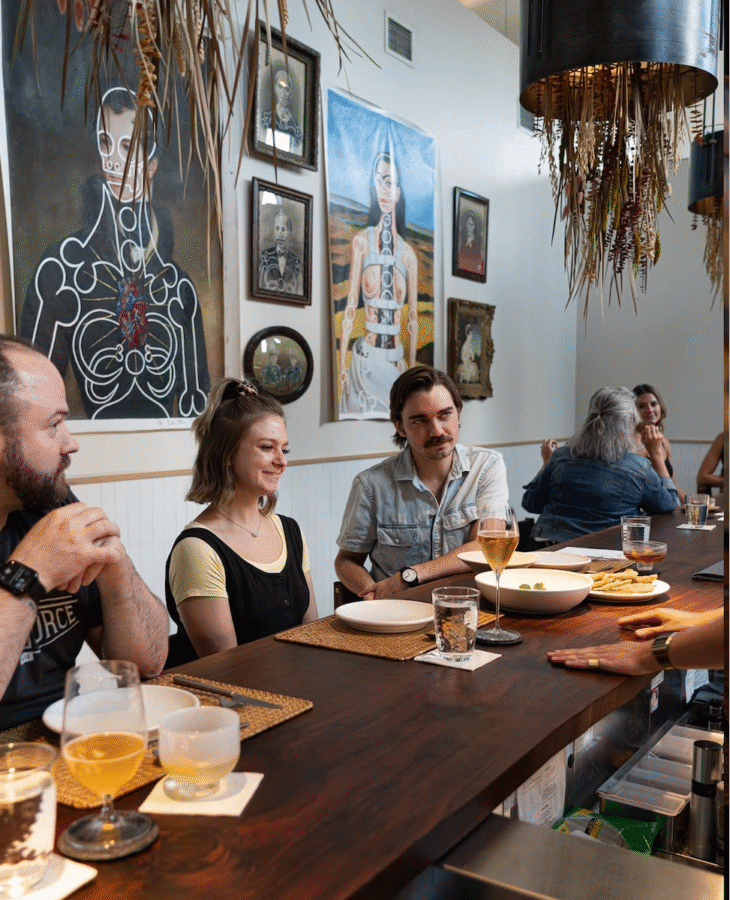 The Rise of Nashville's Vinyl Lounges: 3 to Visit Now + 1 Coming Soon - 2 Three people sit at a bar in a cozy vinyl lounge, enjoying drinks and food while chatting with the bartender. Artwork featuring anatomical figures decorates the wall behind them.
