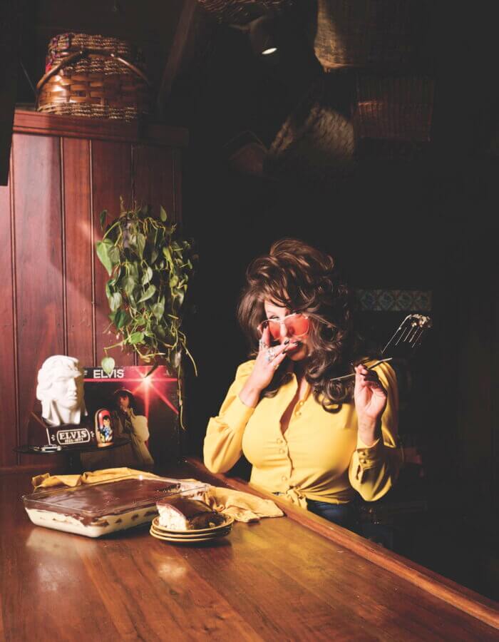 RECIPE: No-Bake Elvis Éclair Cake - 2 A woman in yellow sits at a table with a cake. She's licking her finger and holding a spatula.