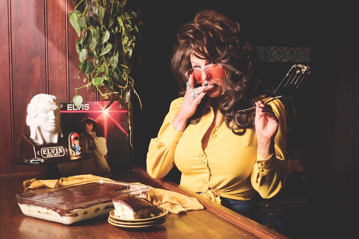 A woman in a yellow blouse holds a spatula and a pair of peaches in front of her face, with an Elvis bust, record, and a dessert on a wooden table.
