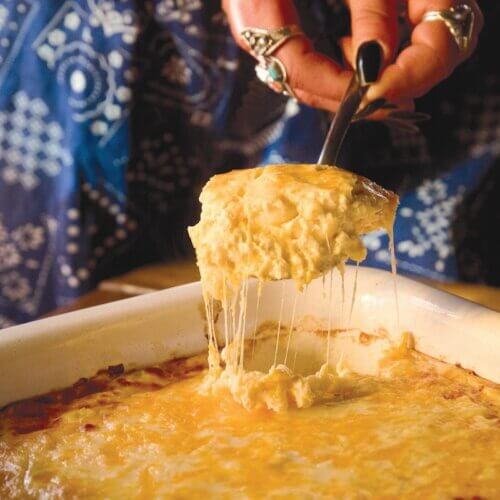 RECIPE: Getchyo' Man Macaroni Cheese - 3 A hand serves a cheesy, golden-brown baked casserole from a rectangular dish, with melted cheese stretching from the serving spoon.