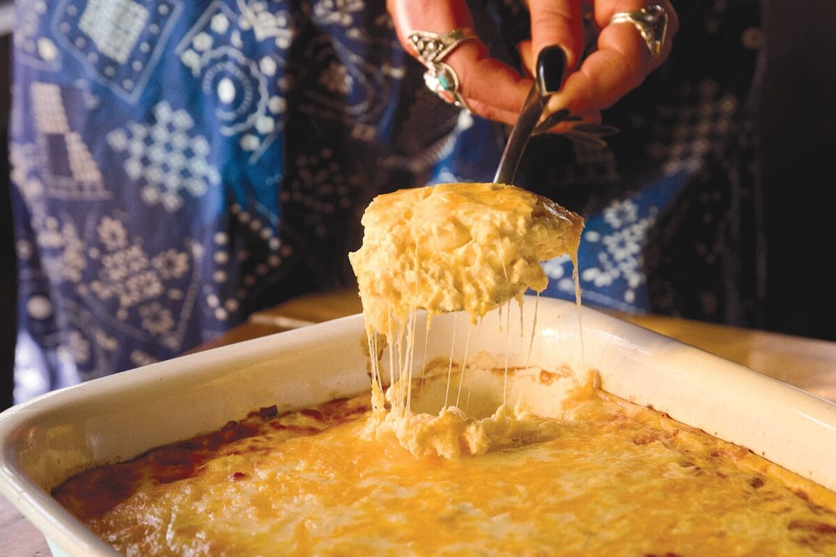 A hand serves a cheesy, golden-brown baked casserole from a rectangular dish, with melted cheese stretching from the serving spoon.