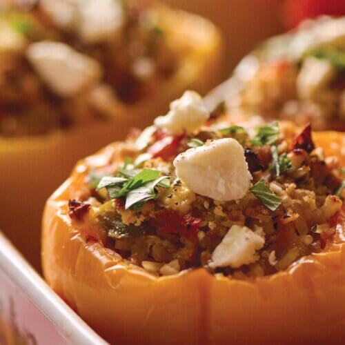 RECIPE: Keep Yo' Man Stuffed Peppers - 3 Close-up of baked yellow bell peppers stuffed with a mixture of grains, vegetables, herbs, and topped with crumbled cheese in a white dish.