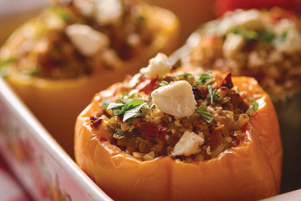 Close-up of baked yellow bell peppers stuffed with a mixture of grains, vegetables, herbs, and topped with crumbled cheese in a white dish.