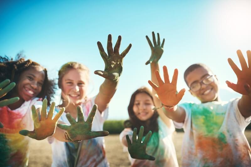 Summer Camp is STILL Where Growth Happens and Core Memories Are Made - 2 Four children outdoors hold up their paint-covered hands toward the camera, smiling in colorful, paint-splattered shirts—capturing joyful summer camp memories on a sunny day.