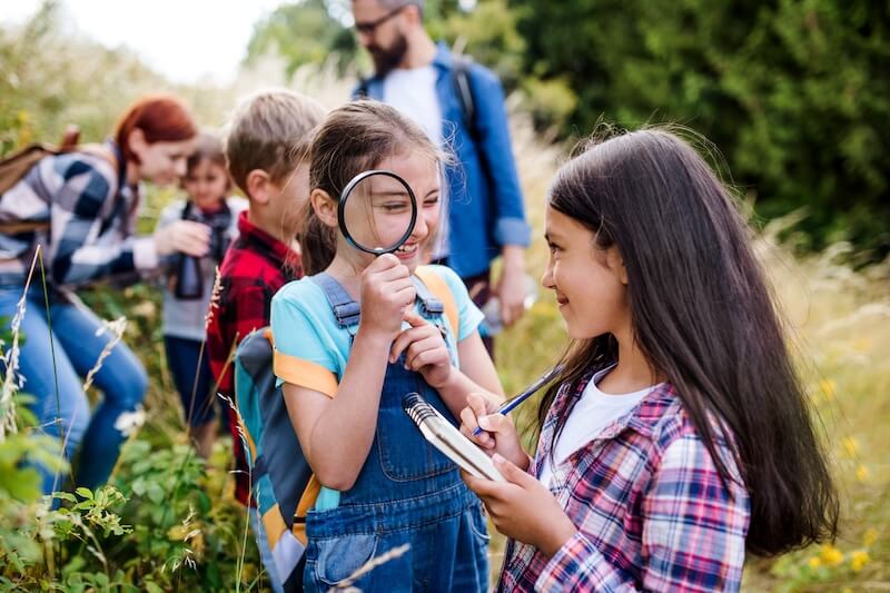 Summer Camp is STILL Where Growth Happens and Core Memories Are Made - 3 Children outdoors, one holding a magnifying glass and another writing in a notebook, with adults in the background capturing summer camp memories during a nature exploration activity.