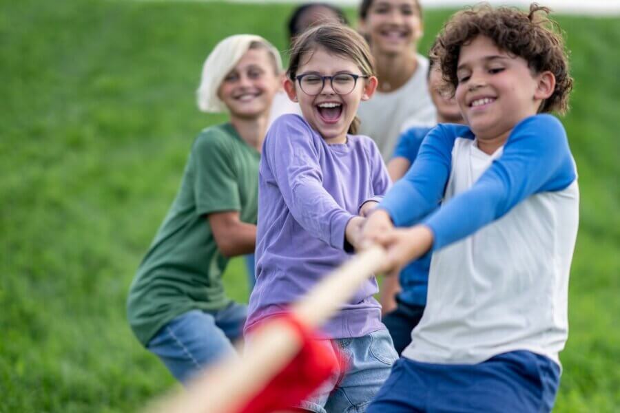 Summer Camp is STILL Where Growth Happens and Core Memories Are Made - 4 A group of children outdoors pulling on a rope in a tug-of-war game, smiling and enjoying the activity, capturing the fun and excitement of summer camp memories.