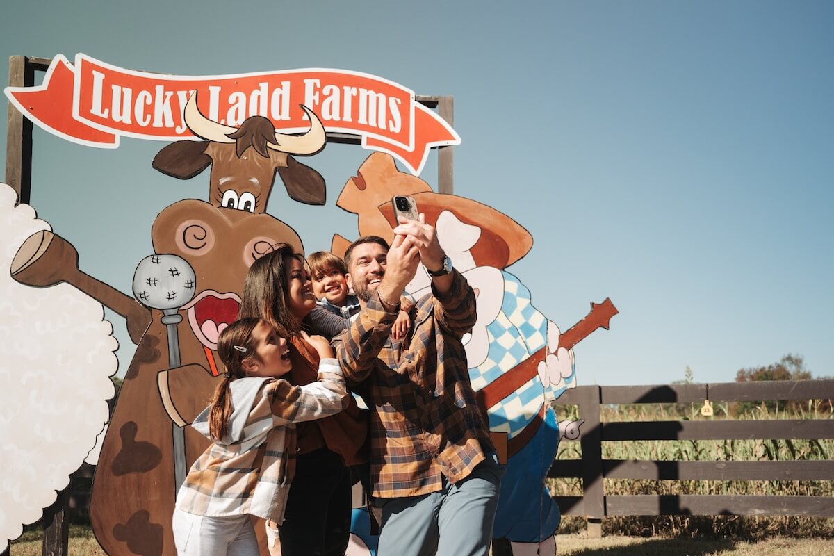 A family of four takes a selfie in front of a "Lucky Ladd Farms" sign featuring cartoon animals on a sunny day—a perfect spot for memorable Tennessee vacations and unforgettable family vacations.