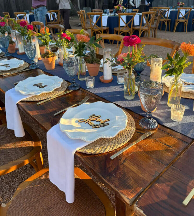 Rustic Meets Romance in This Texas Hill Country Wedding - 8 A rustic wooden table set for a meal with floral centerpieces, candles, woven placemats, and "Thank You" place cards on plates, surrounded by wooden chairs.