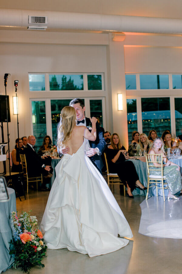 Rustic Meets Romance in This Texas Hill Country Wedding - 16 A bride and groom share their first dance at a Texas Hill Country Wedding reception, surrounded by seated guests in a well-lit venue with large windows.