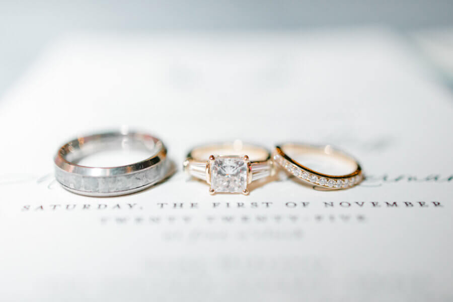 Rustic Meets Romance in This Texas Hill Country Wedding - 3 Three rings—one silver band, one gold diamond band, and a gold ring with a square-cut diamond—rest on a white surface with blurred text behind them, capturing the timeless elegance of a romantic wedding.