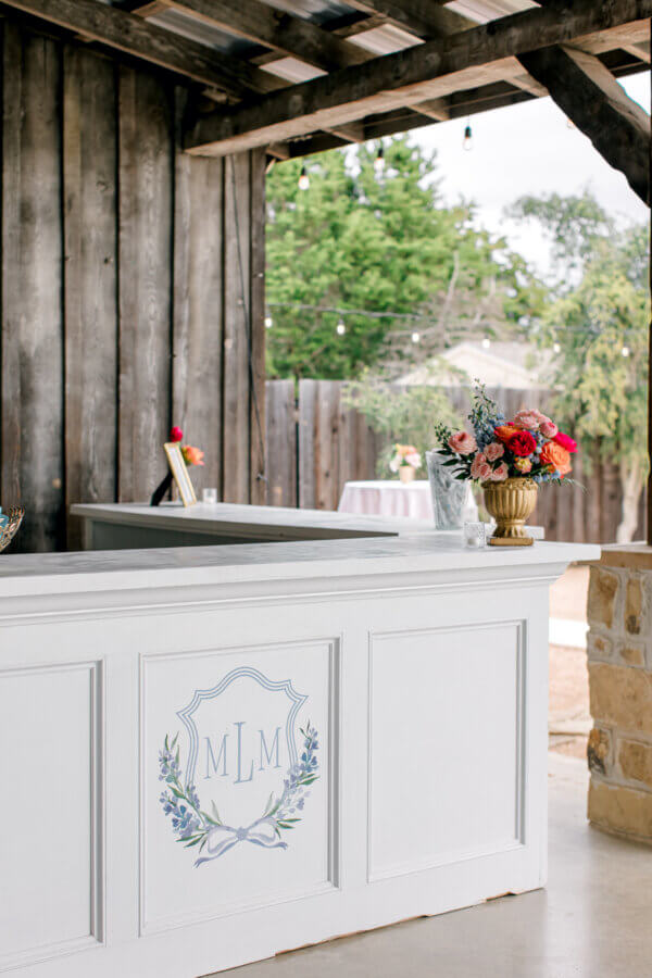 Rustic Meets Romance in This Texas Hill Country Wedding - 6 A white reception bar with floral arrangements and an "MLM" monogram stands under a rustic wooden pavilion, perfect for a romantic wedding in the Texas Hill Country.