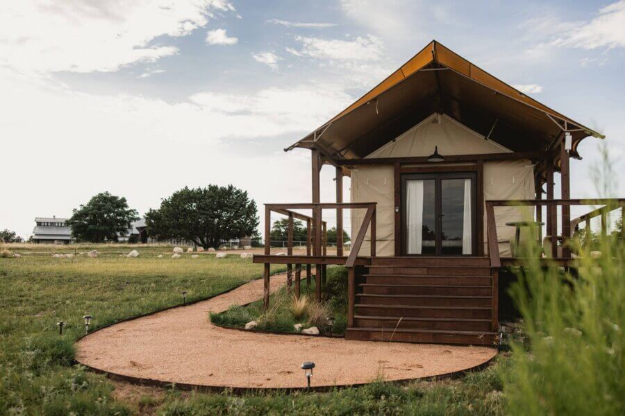 Rustic Meets Romance in This Texas Hill Country Wedding - 5 A small glamping tent cabin with a wooden porch sits on a gravel path in an open grassy field, perfect for a rustic wedding in the Texas Hill Country under a partly cloudy sky.
