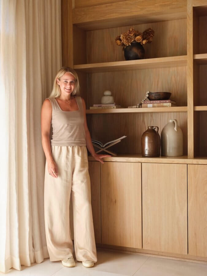 Claire Thompson's Interior Designs Are Soft, Warm, and Endlessly Livable - 2 Claire Thompson stands by a wooden bookshelf adorned with decorative objects, books, and vases, wearing a beige tank top and pants in a softly lit room.