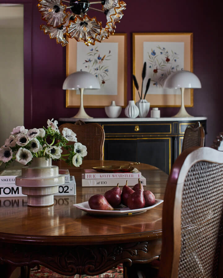Something Old, Something New: How Ellie Christopher Makes Antiques Feel Fresh - 7 A dining room by interior designer Ellie Christopher features a wooden table set with books, a flower vase, and a plate of pears, in front of a dark cabinet and maroon walls adorned with framed art and white lamps.