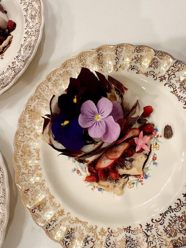 Meet Chef Keshia Hay, the Heart & Soul Behind BōTANIQUE - 4 A decorative plate holds a gourmet dish by Keshia Hay, topped with edible flowers, pomegranate seeds, and thinly sliced vegetables on a creamy base.