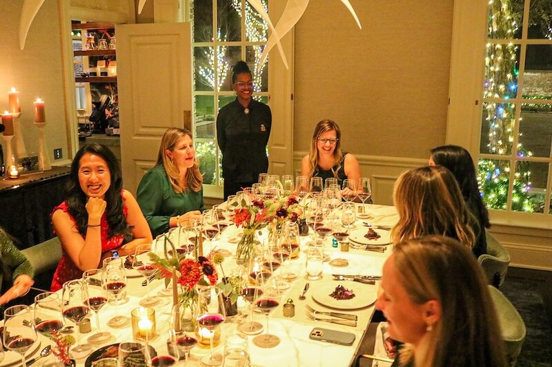 Meet Chef Keshia Hay, the Heart & Soul Behind BōTANIQUE - 3 A group of women sit around a dinner table set with wine glasses and floral centerpieces, while a smiling server stands in the background. The elegant scene reflects Keshia Hay’s signature flair for unforgettable dining experiences.