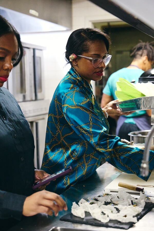 Meet Chef Keshia Hay, the Heart & Soul Behind BōTANIQUE - 5 Two women work in a commercial kitchen; Keshia Hay prepares food at the counter, while the other arranges items on a tray and holds her phone. In the background, a man works, focused on his task.