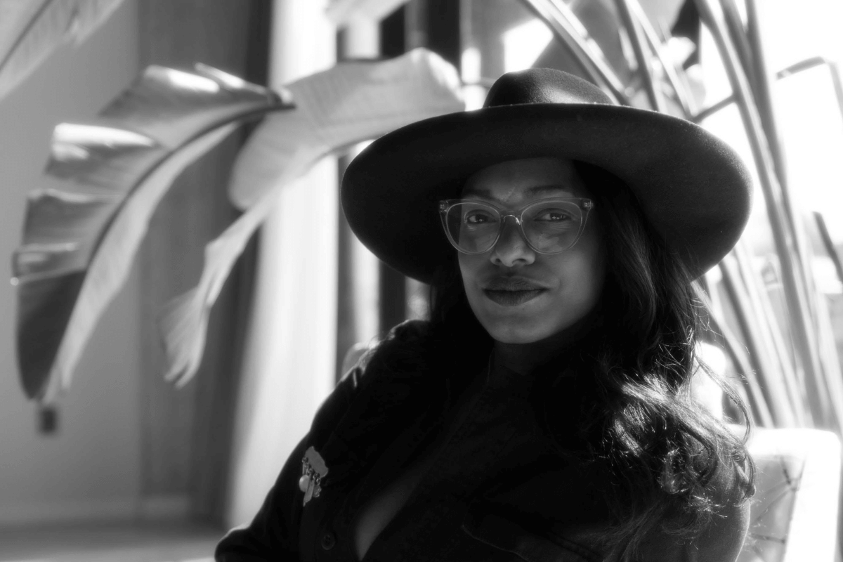 Black and white photo of Keshia Hay, FACE framed by long hair and glasses, wearing a wide-brimmed hat and dark shirt, sitting indoors near large plant leaves.
