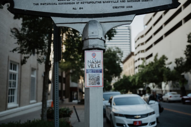 She’s Putting Nashville’s Hidden History on the Map - 4 A metal signpost with a "Nashville Sites" sign and QR code stands on a city sidewalk, highlighting locations curated by Dr. Mary Ellen Pethel, with parked cars and buildings visible in the background.