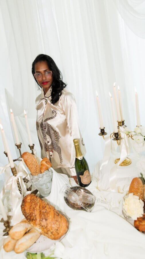 Meet Chef Keshia Hay, the Heart & Soul Behind BōTANIQUE - 2 A woman in satin pajamas, stands behind a table set with bread, champagne, candles, and white flowers against a white draped background.
