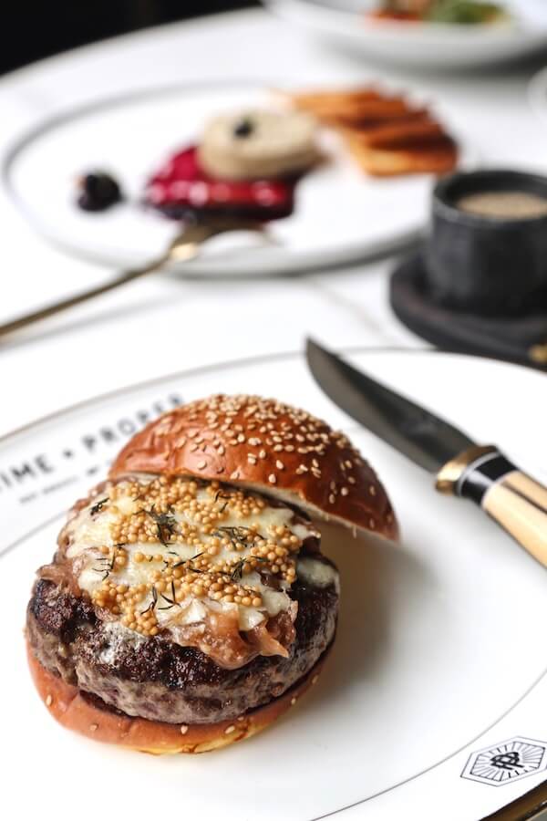 20 Epic Spots to Grab Dinner at the Bar in Nashville - 9 A cheeseburger with a sesame seed bun sits on a white plate next to a knife, perfect for dinner at a lively Nashville bar, with blurred dessert and coffee in the background.