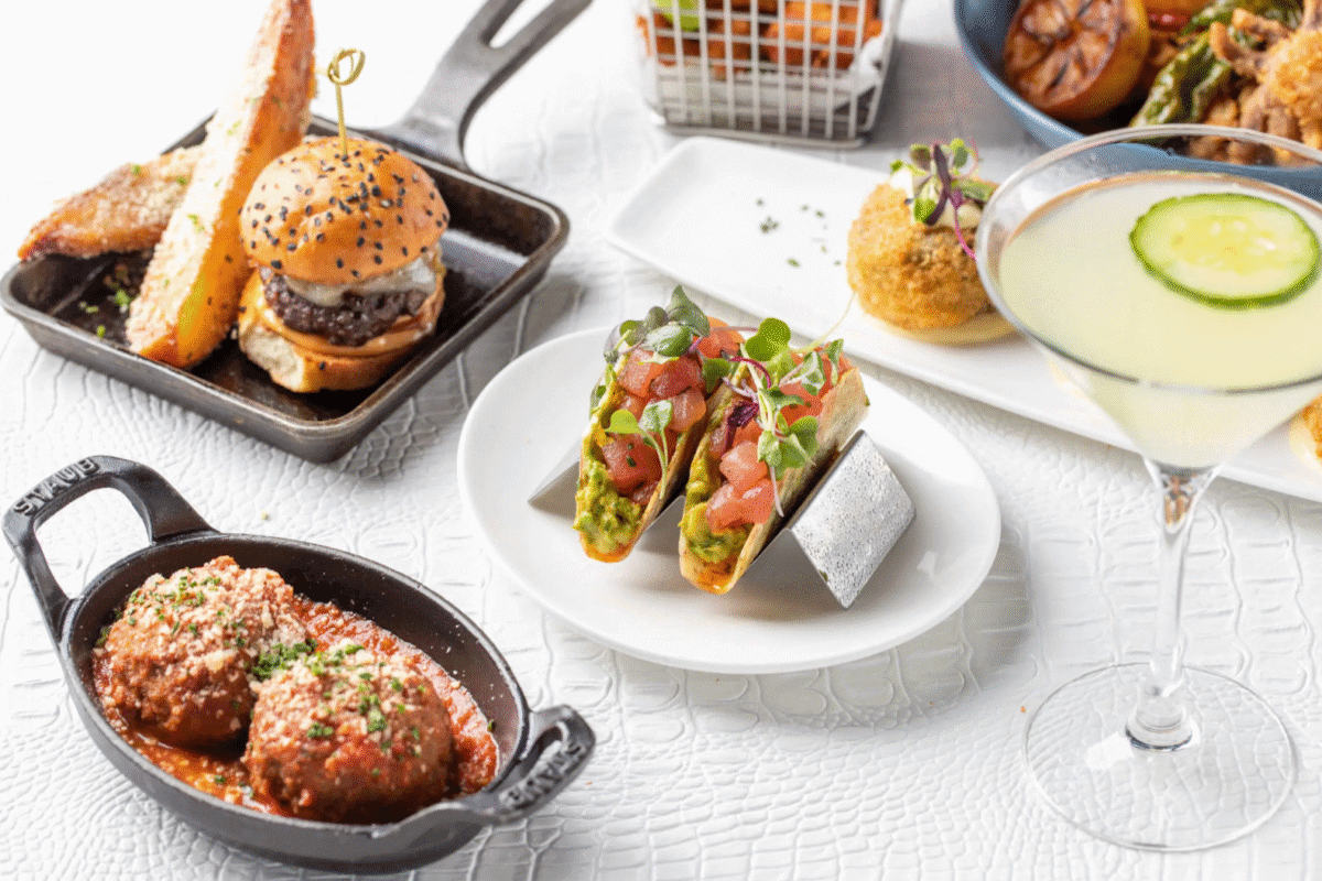 Assorted appetizers—perfect for dinner at the bar—including sliders, tacos, meatballs, croquettes, and a cucumber cocktail are artfully arranged on a white table.