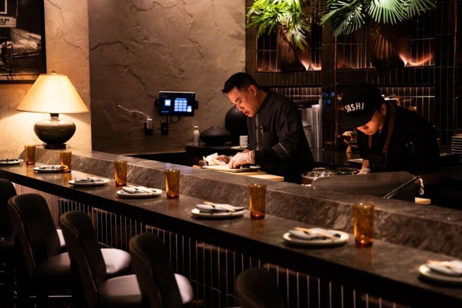 20 Epic Spots to Grab Dinner at the Bar in Nashville - 10 Two chefs work behind a dimly lit sushi bar in Nashville, set with plates, chopsticks, and amber glasses; modern decor and plants complete the inviting Dinner at the Bar atmosphere.