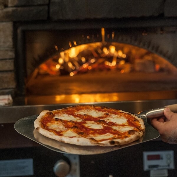 20 Epic Spots to Grab Dinner at the Bar in Nashville - 7 A freshly baked pizza on a metal peel is being removed from a wood-fired oven with visible flames inside, making it the perfect choice for dinner at the bar.