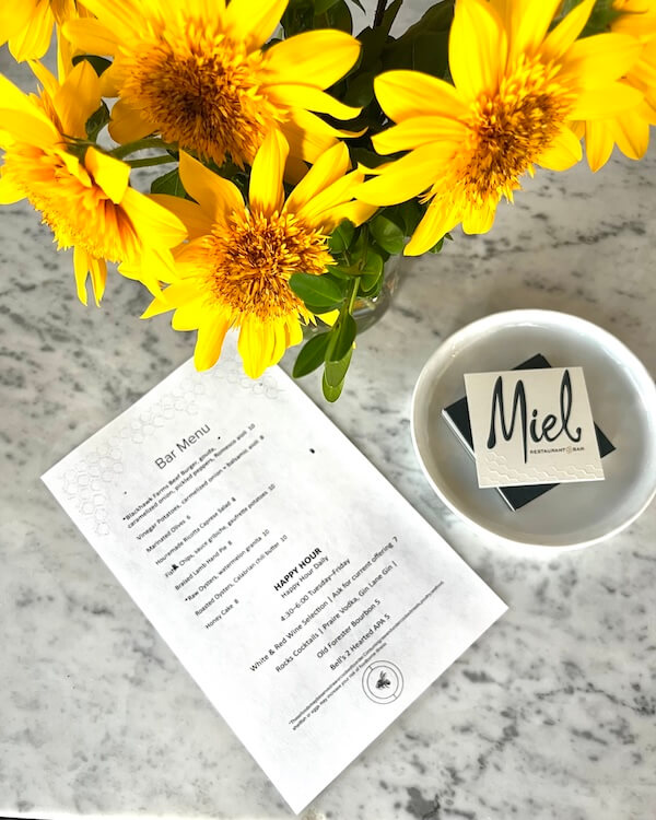 20 Epic Spots to Grab Dinner at the Bar in Nashville - 8 A marble table displays a bouquet of yellow flowers, a bar menu, and a dish holding coasters with "Miel" printed on them—inviting you to enjoy dinner at the bar.
