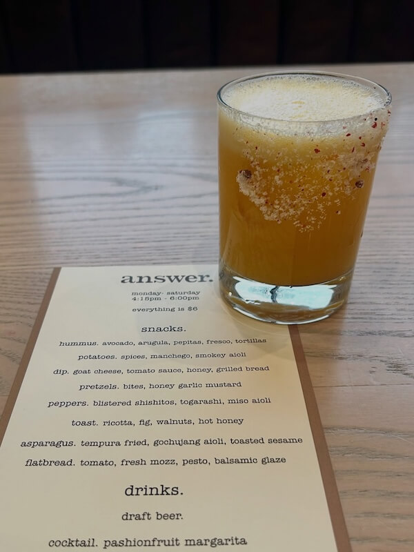 20 Epic Spots to Grab Dinner at the Bar in Nashville - 2 A passionfruit margarita with a salted rim sits on a wooden table next to a printed restaurant menu listing snacks, flatbread, drinks, and cocktails—perfect for enjoying dinner at the bar.