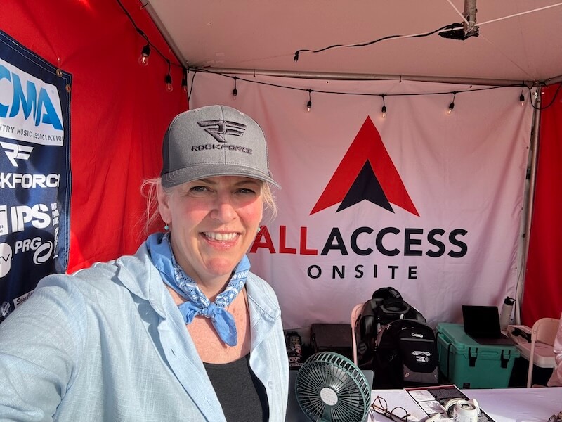 She's Building a Support Network for Nashville's Live Music Community - 2 Kara Kemp, wearing a cap and blue bandana, stands in front of an "ALL ACCESS ONSITE" banner inside a tent filled with event equipment and a small fan.
