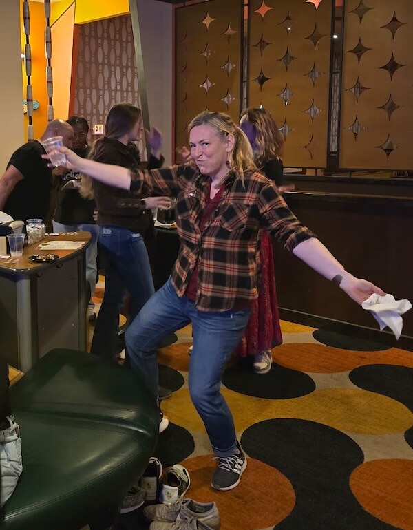 She's Building a Support Network for Nashville's Live Music Community - 4 Kara Kemp, in a plaid shirt and jeans, poses with arms outstretched holding a drink at an indoor social gathering, with others mingling in the background.