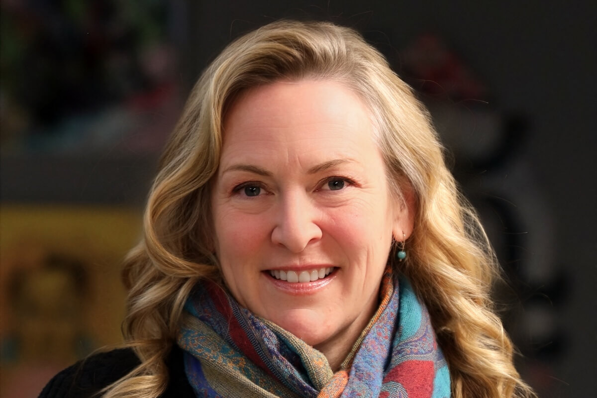 Kara Kemp, a woman with long blonde hair, wears a colorful scarf and a black top as she smiles warmly at the camera against a blurred background.