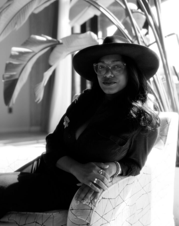 Meet Chef Keshia Hay, the Heart & Soul Behind BōTANIQUE - 6 A woman wearing glasses and a wide-brimmed hat sits on a patterned chair, posing with her hands folded, in a well-lit room with large leafy plants in the background.