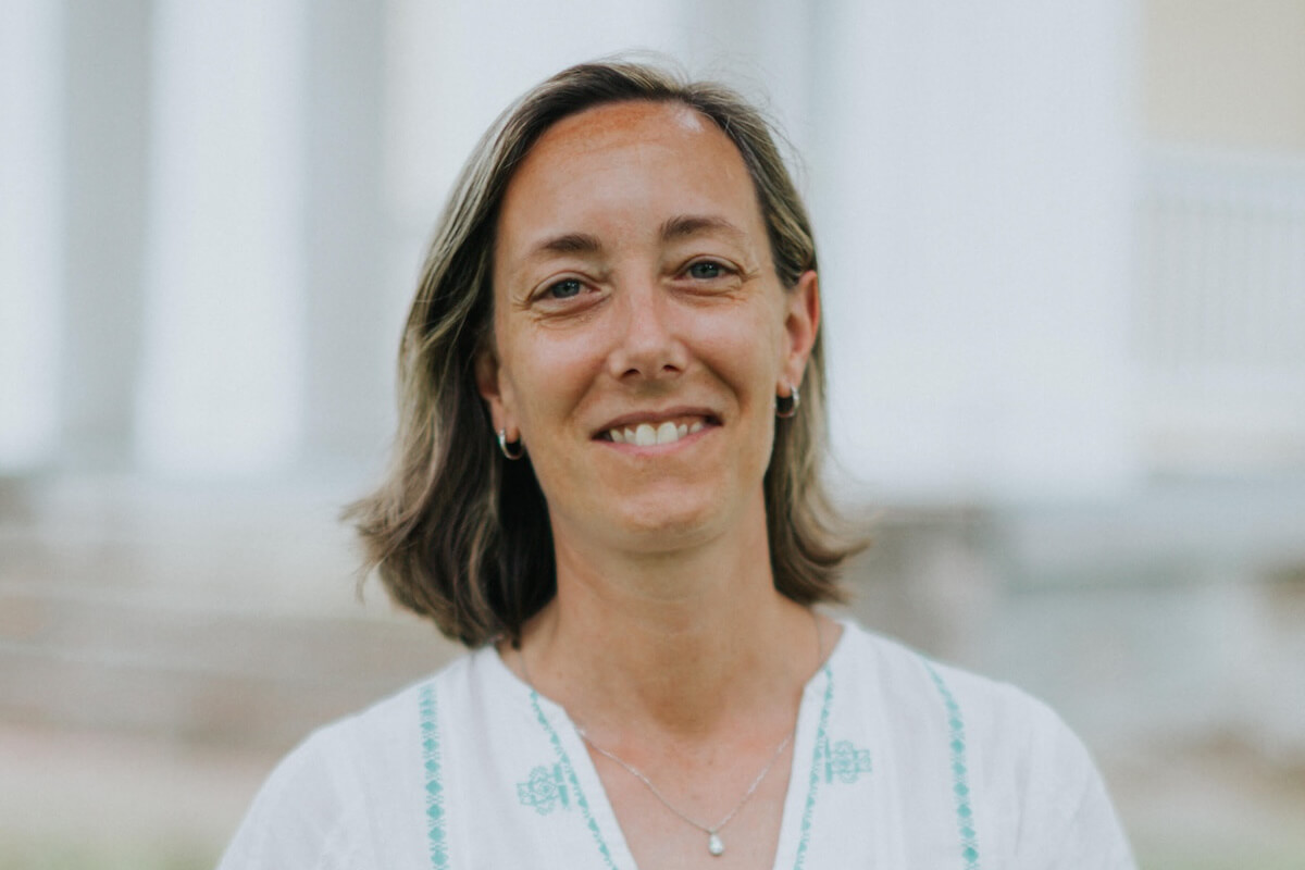 Dr. Mary Ellen Pethel, a woman with shoulder-length brown hair, wearing a white top with blue embroidery, smiles at the camera in an outdoor setting.