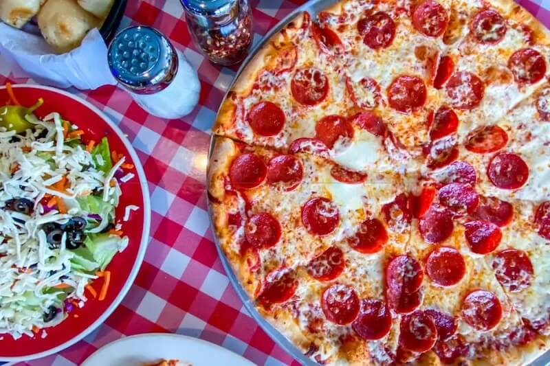 A pepperoni pizza on a metal tray, a plate of salad with shredded cheese, salt and pepper shakers, and rolls on a red and white checkered tablecloth—perfect for sampling the flavors of new restaurants opening in Nashville in February 2026.