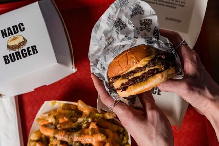 A person holds a double cheeseburger wrapped in paper above a tray of loaded fries, with a NADC Burger takeout box in the background.