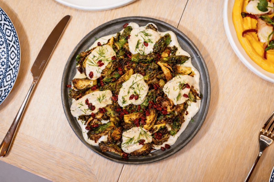 A grey plate with roasted Brussels sprouts topped with a creamy white sauce, herbs, and red berries, styled as served in new Nashville restaurants, sits on a light wooden table with utensils nearby.