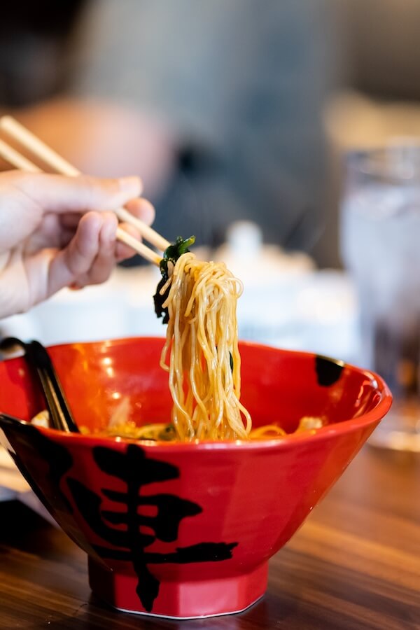 A hand holding chopsticks lifts noodles from a red bowl of ramen on a wooden table, capturing the essence of Nashville restaurants set to debut in February 2026.