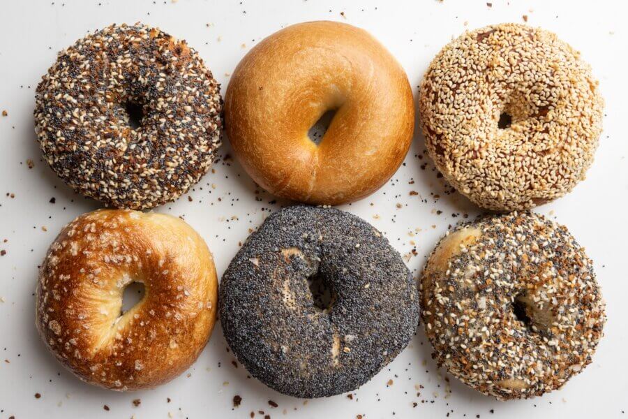 Six assorted bagels with poppy seeds, sesame seeds, and everything seasoning, arranged on a white surface with scattered crumbs.