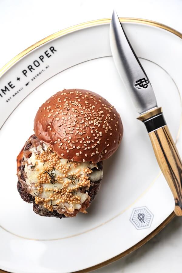 A cheeseburger with a sesame seed bun is served on a white plate with "TIME + PROPER" written on it, next to a steak knife with a wooden handle—a standout among new Nashville restaurants opening February 2026.