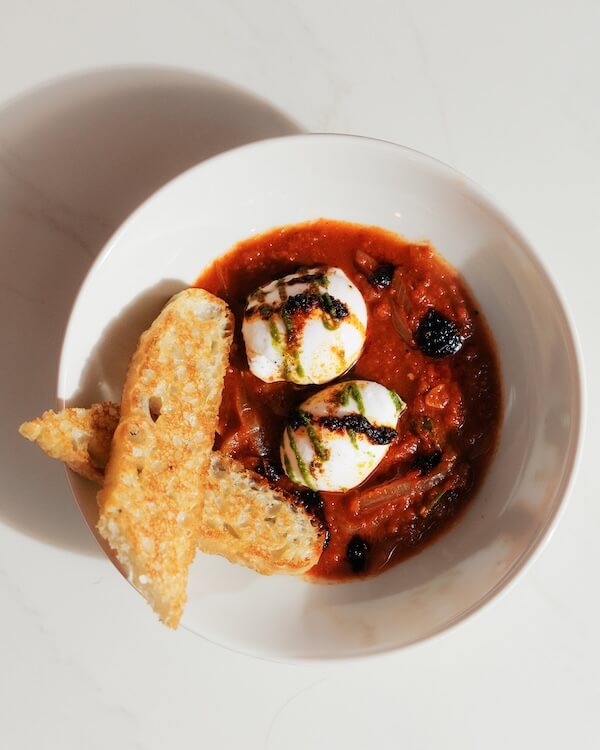 A white bowl with two poached eggs in tomato sauce, garnished with green herb oil and served with toasted bread—a standout dish inspired by the flavors at new Nashville restaurants.