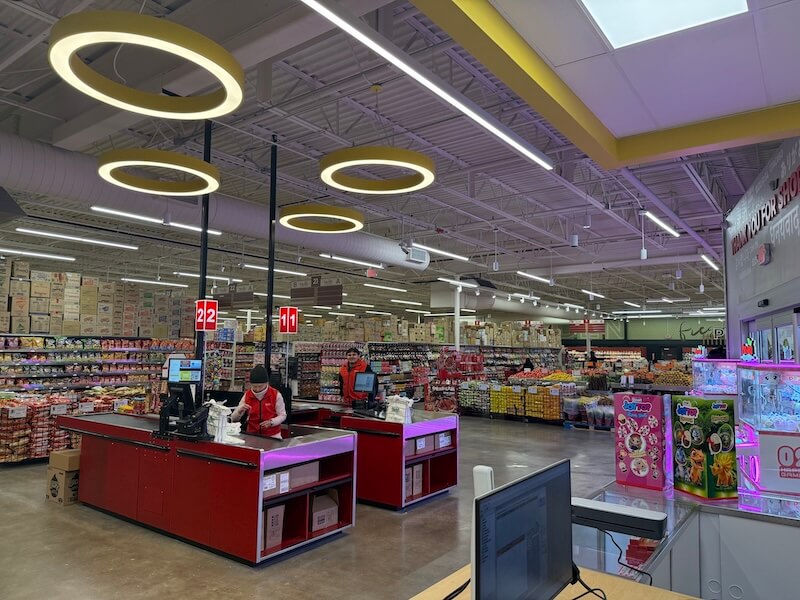 9 New Nashville Retail Spots to Explore - 8 Two supermarket cashiers in red uniforms check out customers at red registers, with store shelves, produce, and bright circular ceiling lights visible—a lively scene at one of the newest Nashville retail spots.