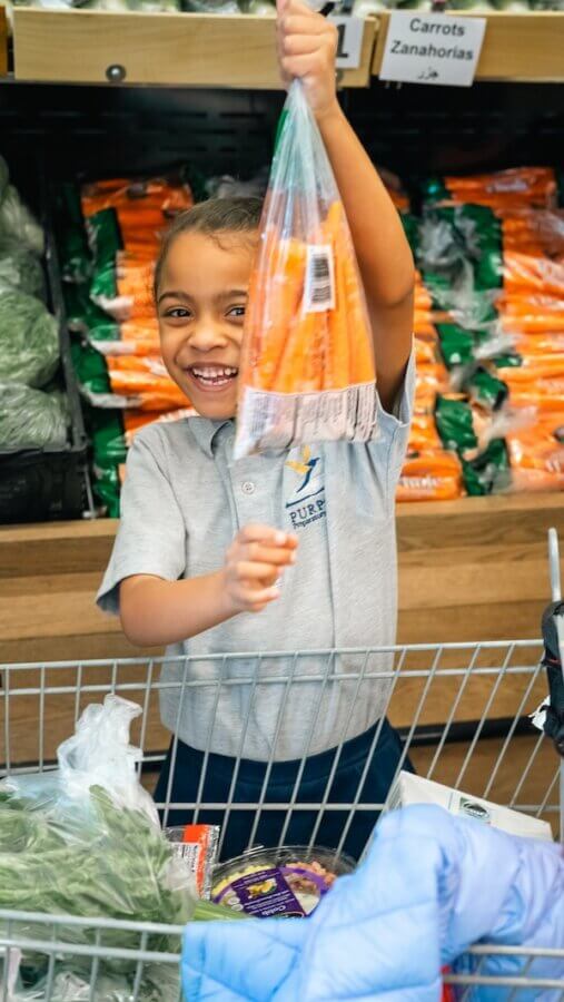 Turnip Truck & 'The Store' Are Rounding Up for Neighbors in Need - 4 A child stands in the store, smiling and holding up a bag of carrots above a shopping cart filled with groceries, looking like they just hopped off the turnip truck.